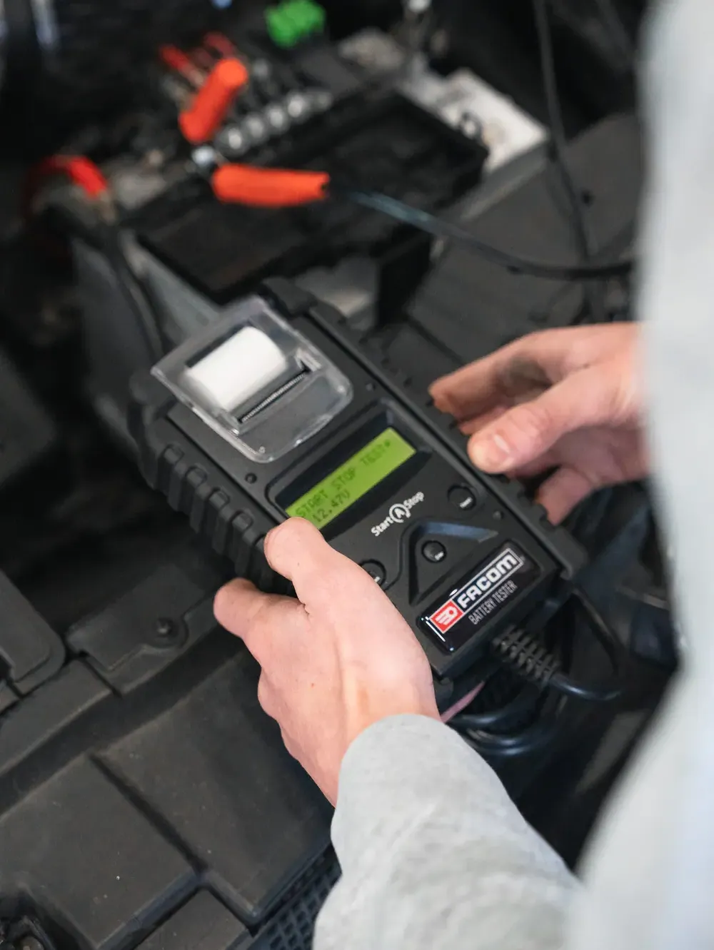 Deux mains tiennent un testeur de batterie Facom au-dessus du compartiment moteur