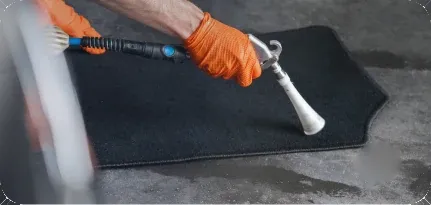 Un gant orange manie un outil de nettoyage sur un tapis de voiture noir